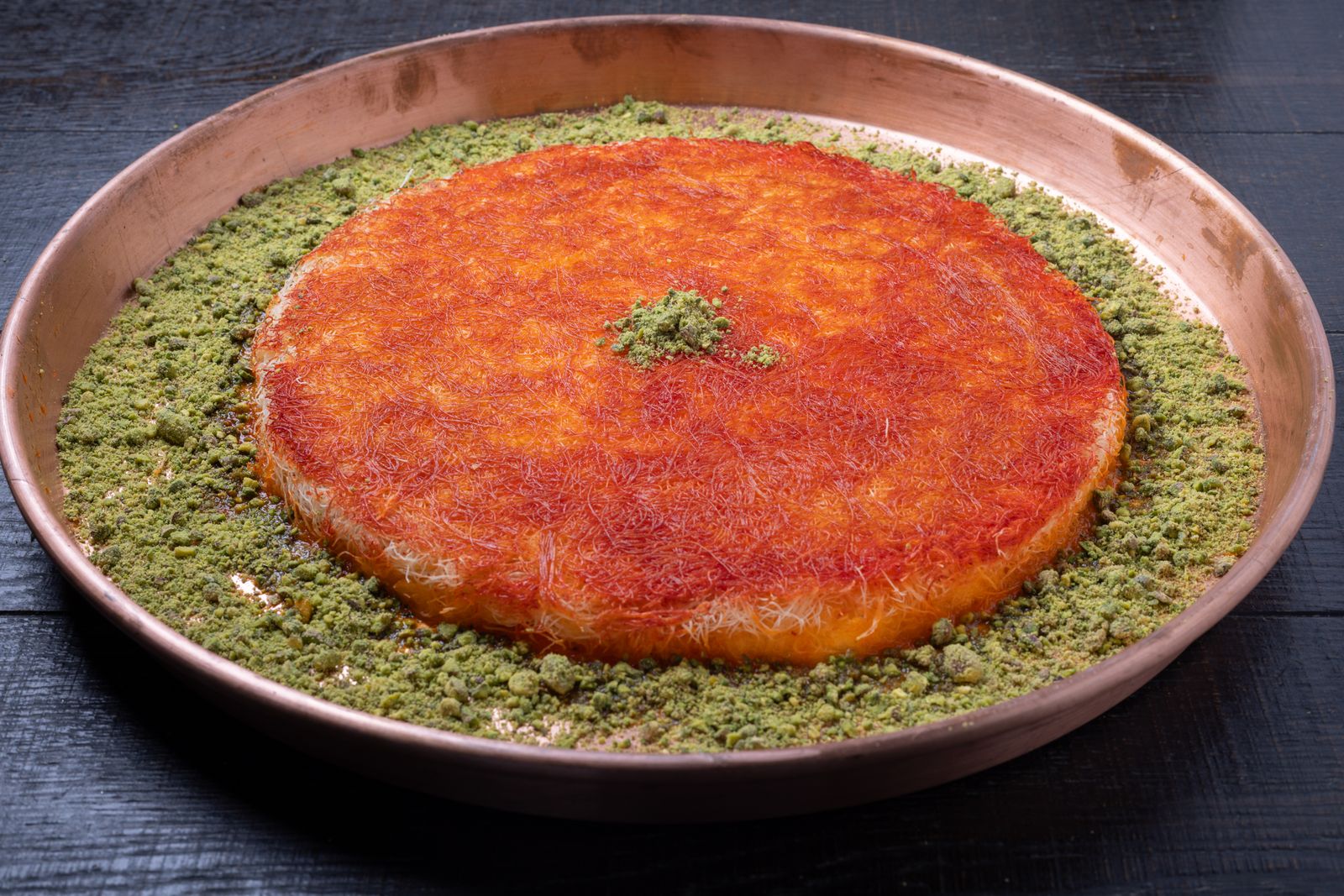 A round copper tray of golden kunafa ringed with ground pistachio